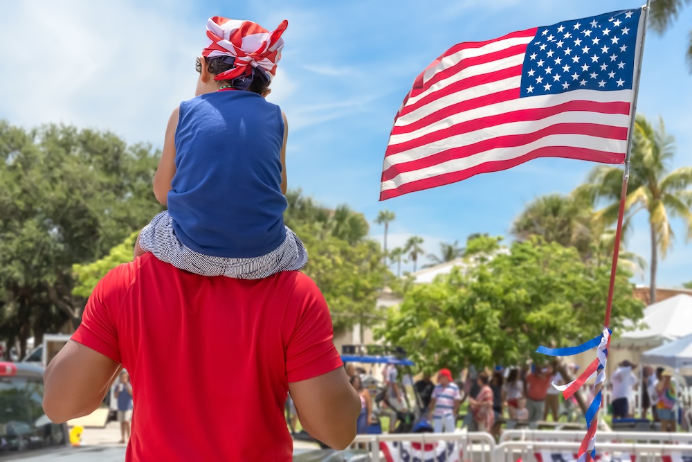 The son wearing a flag bandana on top of his dad's shoulder passing by the American flag.