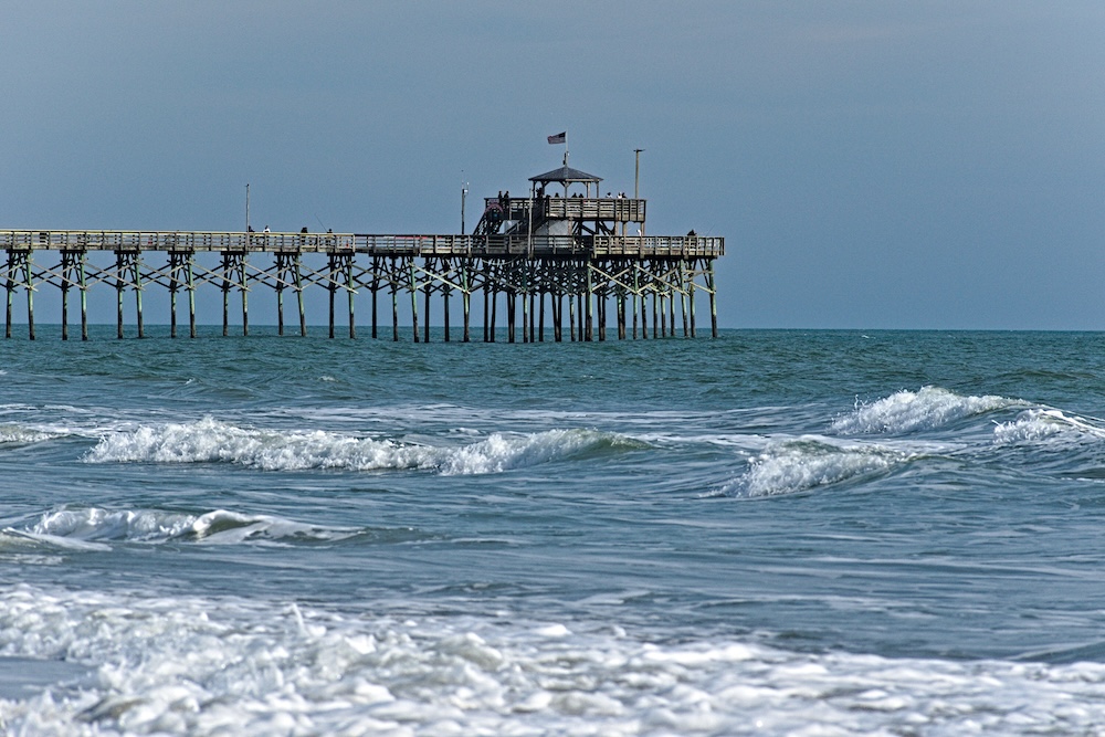 A long wooden pier with a small observation deck and an American flag stands over the blue ocean, with white-capped waves rolling towards the beach.