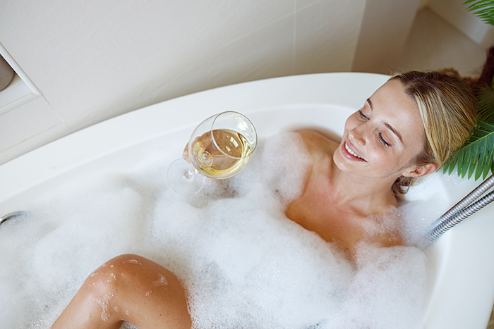 Pretty young Caucasian female relaxing in bath with bubbles and sipping white wine. Side view.