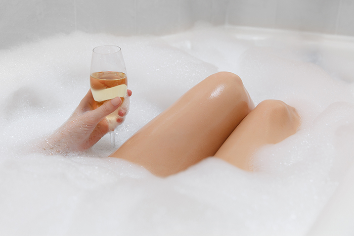 Close up photo of young sexy woman with straight naked body and clean legs lying and relaxing in white foam bath tub with candles around in light bathroom, drink alcohol from wine glass indoors