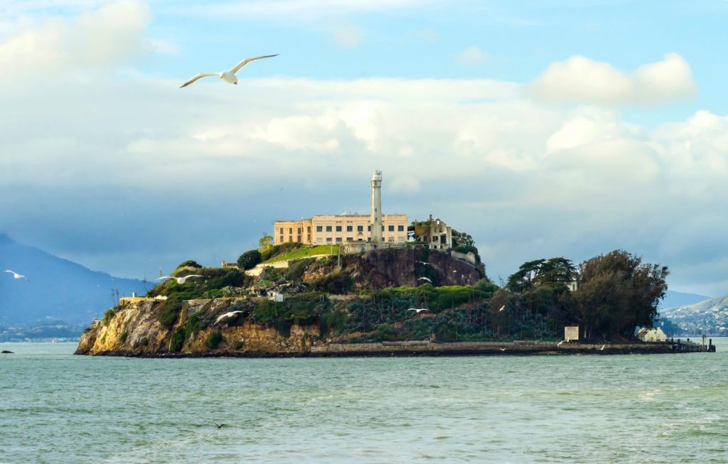 The Alcatraz Penitentiary, now a museum, in San Francisco, California, United States of America. A view of the island, the lighthouse, prison buildings and the San Francisco Bay from the coast on a sunny day.