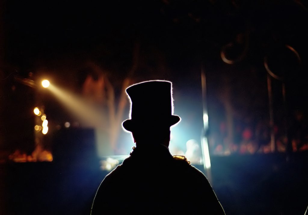 Performer with a hat on stannding on a dark stage facing silhouette of audience members.