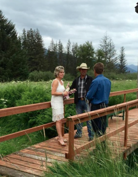 A man in a cowboy hat officiates a wedding ceremony between a bride and groom standing on a wooden pedestrian bridge on the property of the lodge