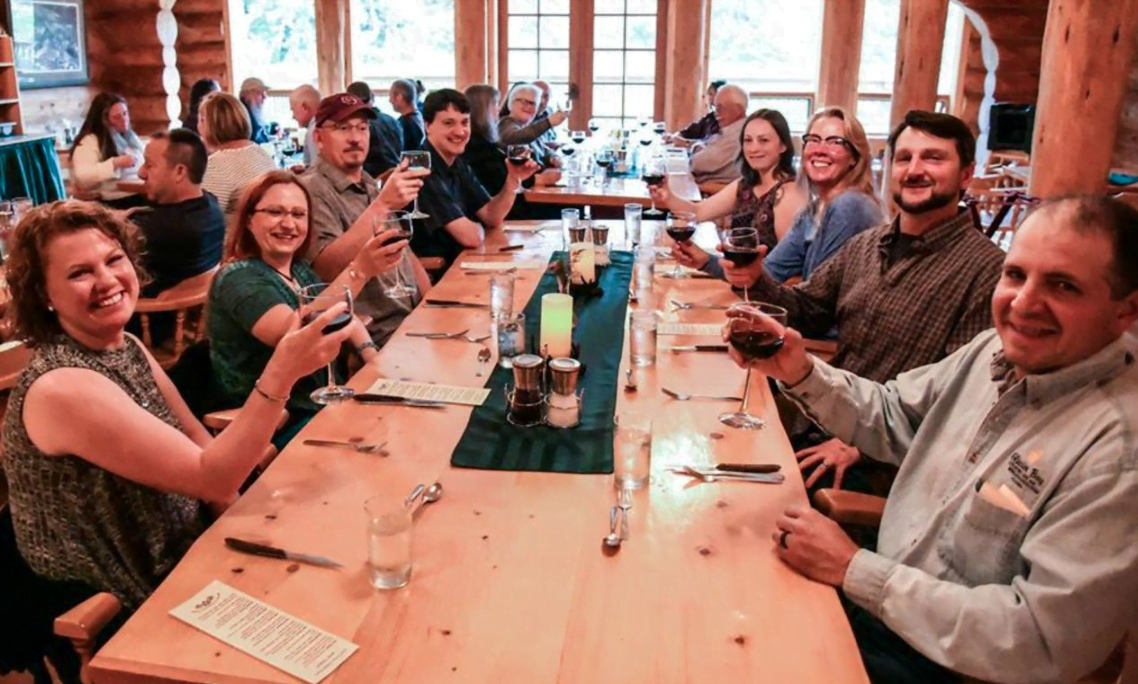 A group of men and women sit at a long table in the lodge's dining room holding up glasses of wine and smiling at the camera