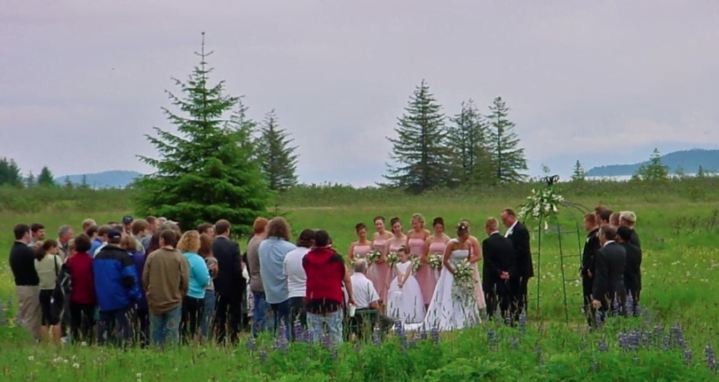 A wedding ceremony takes place in a large grassy meadow on the property of the lodge. The bridal party stands up front featuring bridesmaids in pink dresses, a flower girl, the bride, the groom, the officiant, and groomsmen in black suits. A large audience stands and watches.