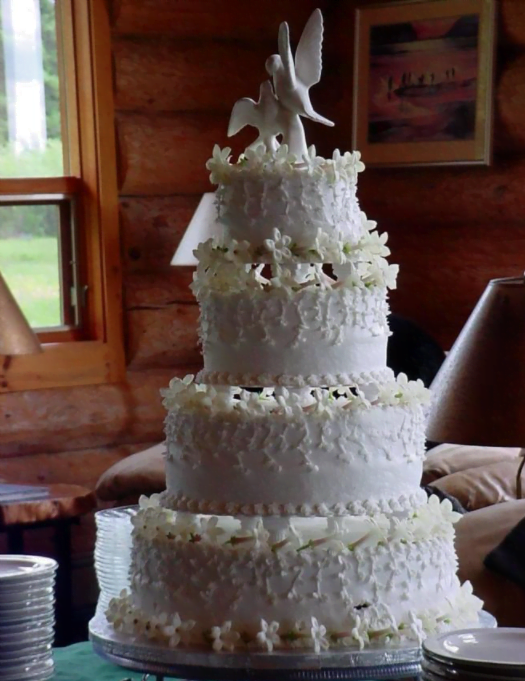 A four tiered wedding cake with decorative white flowers and a cake topper with two white doves sits on a green table in the lobby of the lodge
