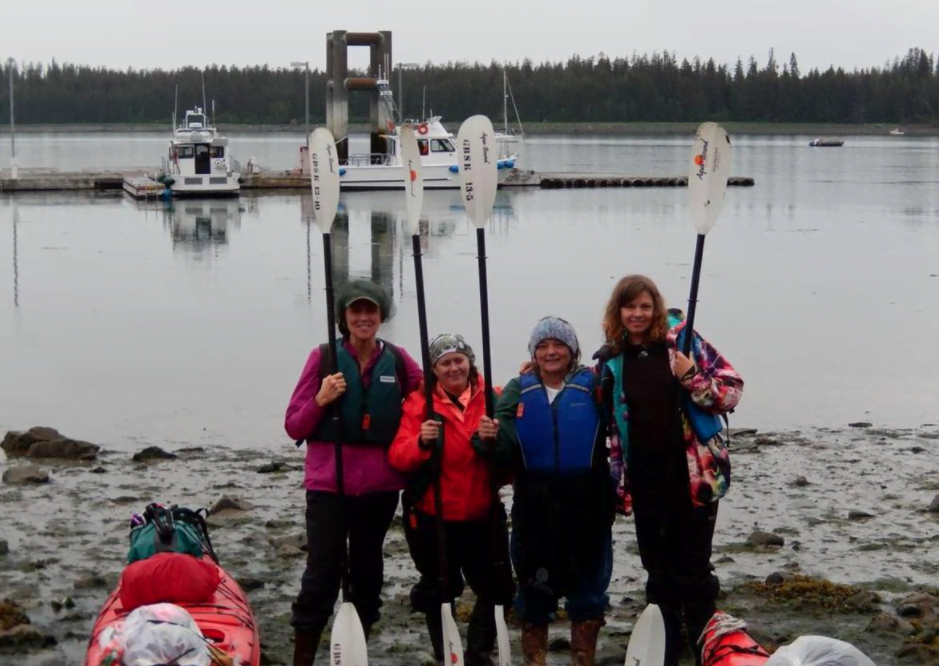 A group of four women in thermal clothes each hold a single kayak paddle upright while standing on a beach. A large lake with several fishing boats is behind them