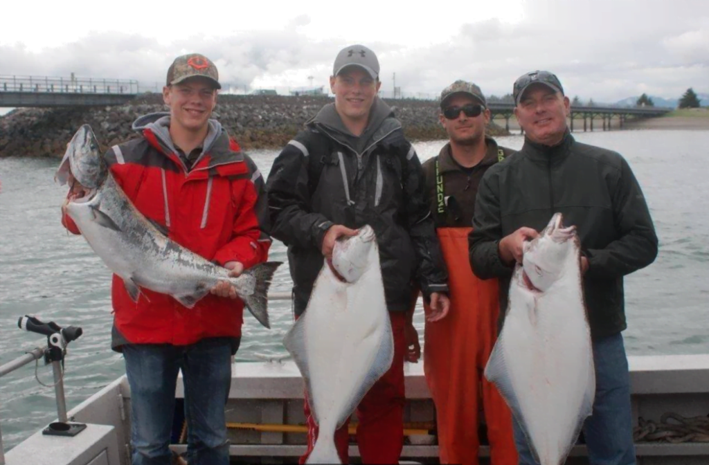 A group of men smile and hold up the halibut and salmon they caught while on the back of a fishing boat