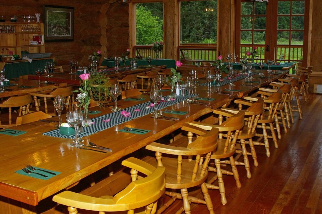 A dining room table in the lodge is set up for a formal dinner and decorated with pink flowers in glass vases, empty wine glasses, and green linens
