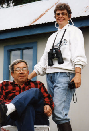 Bear Track Inn owner Jane wearing glasses and binoculars stands next to her husband John who is seated in a chair wearing a red flannel shirt