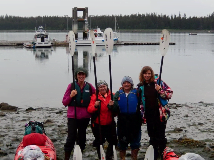 A group of four women in thermal clothes each hold a single kayak paddle upright while standing on a beach. A large lake with several fishing boats is behind them