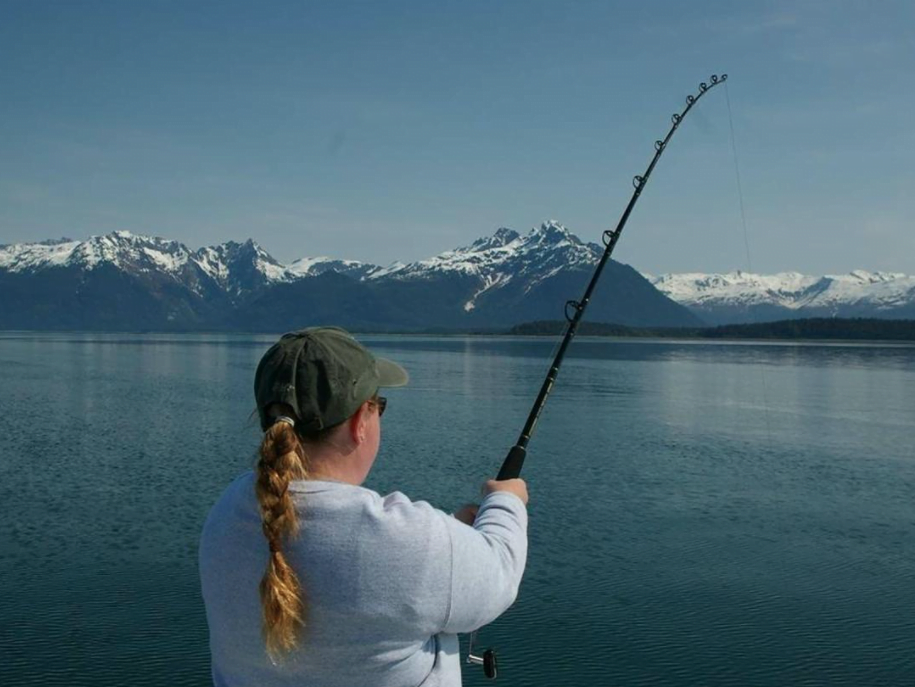 A woman with her back turned to the camera holds a fishing rod cast into the ocean with a view of snow covered mountains in the background