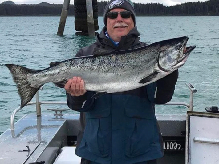 A man in fishing gear grins as he holds up a large silver salmon he caught while on the back of a fishing boat