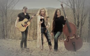 The Mountain Gypsies band pictured here. There are 3 people in a wintery, muted toned, wooded area. The man is on the far left with a guitar. There is a young adult lady in the middle holding a fiddle, and a woman with a stand up bass on the far right.