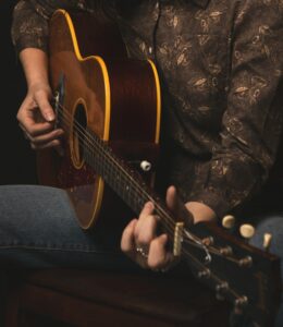 Hand strumming an acoustic guitar. Guitar is at an angle. Person's body is all you see. They are wearing a brown floral shirt and jeans.