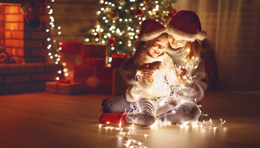 mother holds daughter while both gleefully look down at a string of christmas lights that rests upon their laps