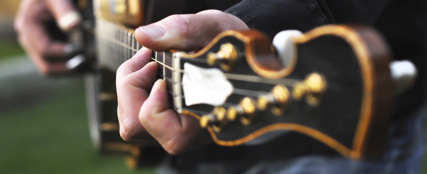 A closeup on a man's left hand playing a Banjo. The Banjo is out of focus except for the neck where his hands are playing a chord. The Banjo hangs from a leather strap around his body. His head is not shown.