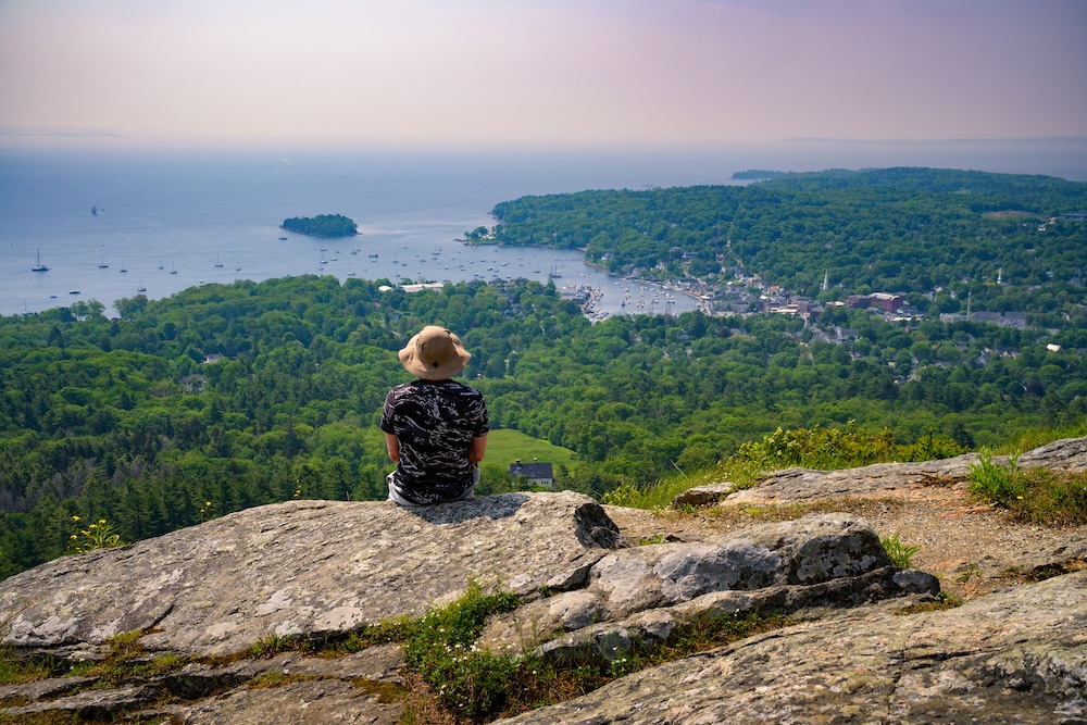 A hiker sits on a rocky ledge at the summit of Mt. Battie in Camden Hills State Park, overlooking Camden Harbor and Penobscot Bay in Maine.
