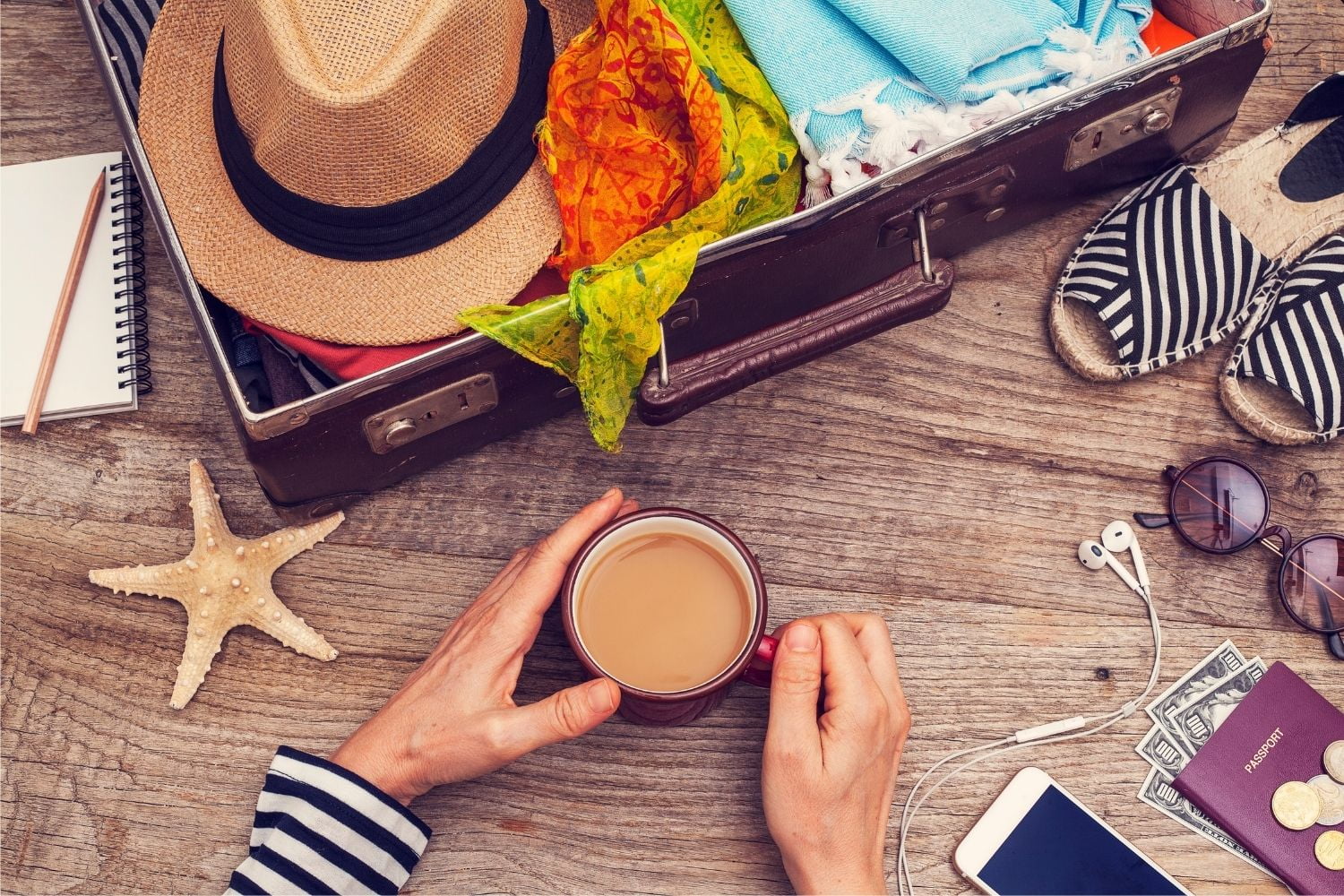 Suitecase filled with items for a vacation. Woman holding coffee cups in her hands.