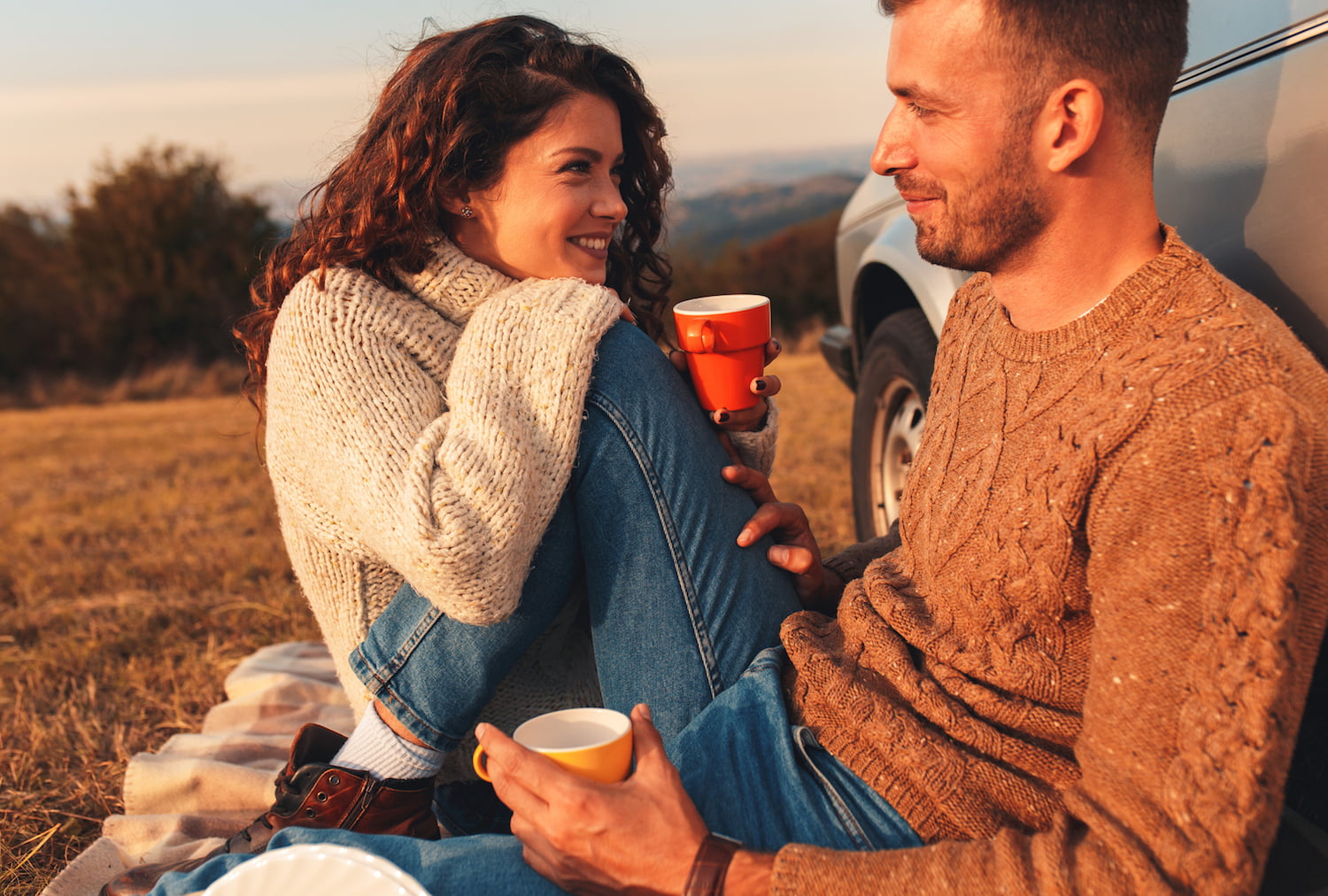 Beautiful young couple enjoying picnic time on the sunset. They drinking tea and sitting in a meadow leaning against a old fashioned car.