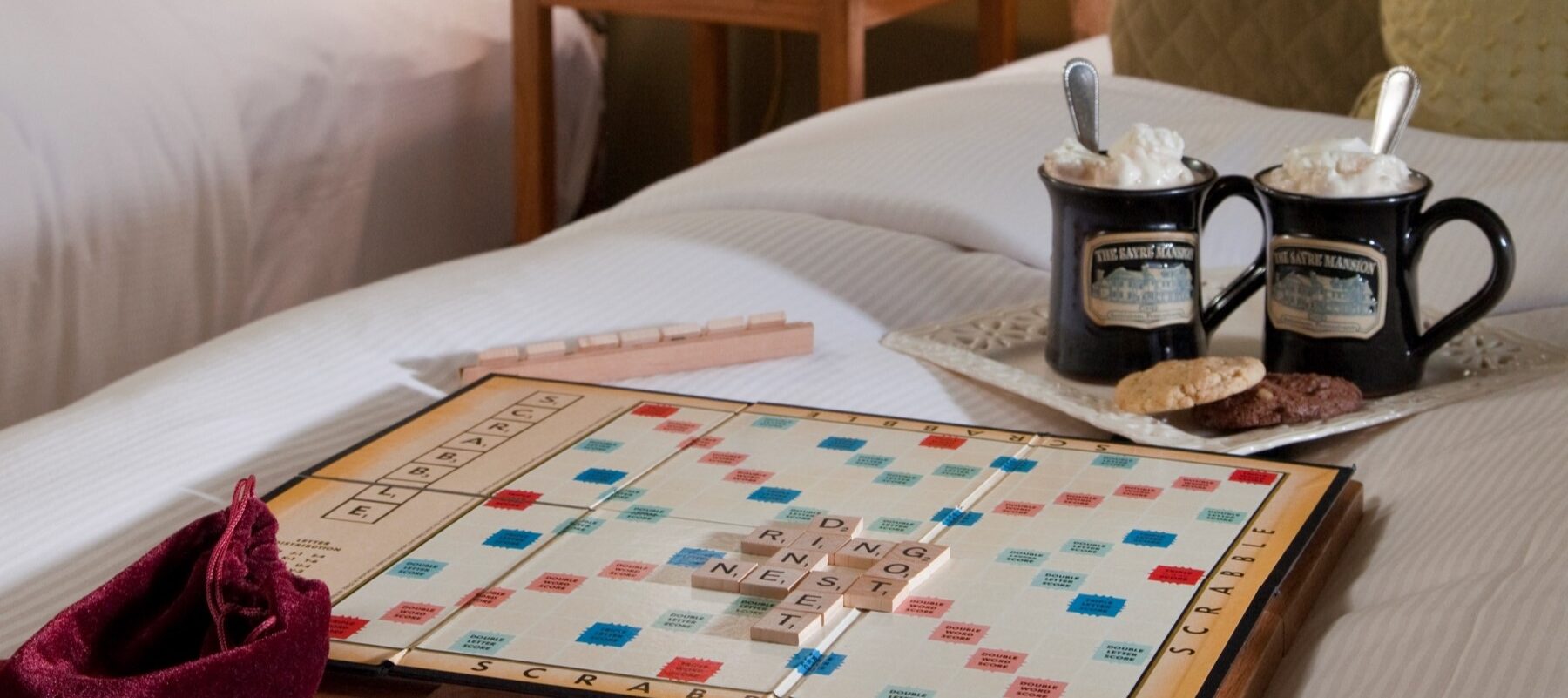 two brown mugs with "the sayre mansion" and a pair of cookies sit on top of a plate resting on a bed, behind a scrabble board