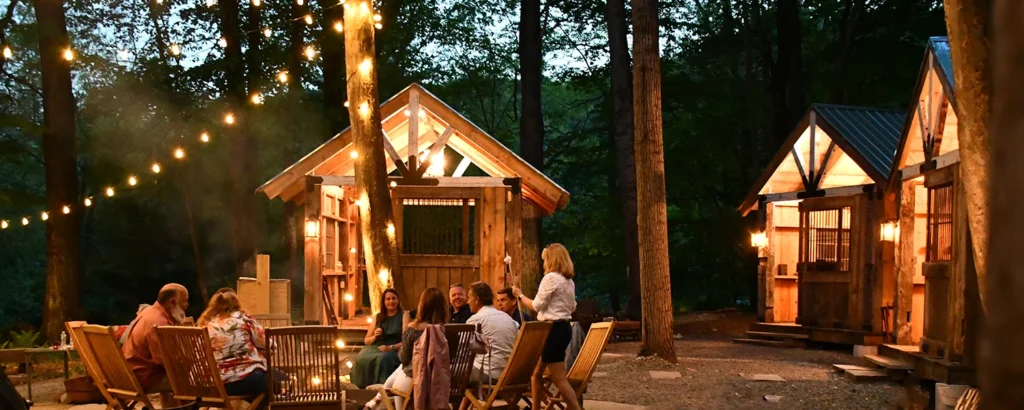 a group of people sits around a campfire under string lights at sunset in hawley, pa