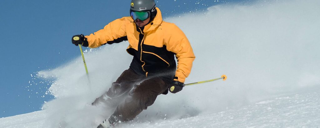 Man skiing down a mountain with a yellow jacket and snow kicking up behind him.