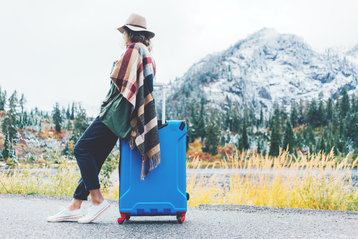 Traveling woman wearing poncho and hat sitting on her bright blue suitcase near stunning mountain wilderness. Wanderlust and boho style (Traveling woman wearing poncho and hat sitting on her bright blue suitcase near stunning mountain wilderness. Wand